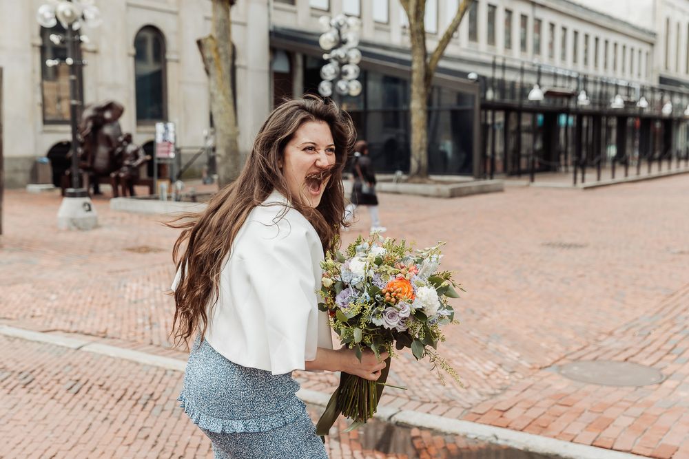 Joyful woman with long hair holding a colorful bouquet on a cobblestone street, wearing a white blouse and blue skirt.