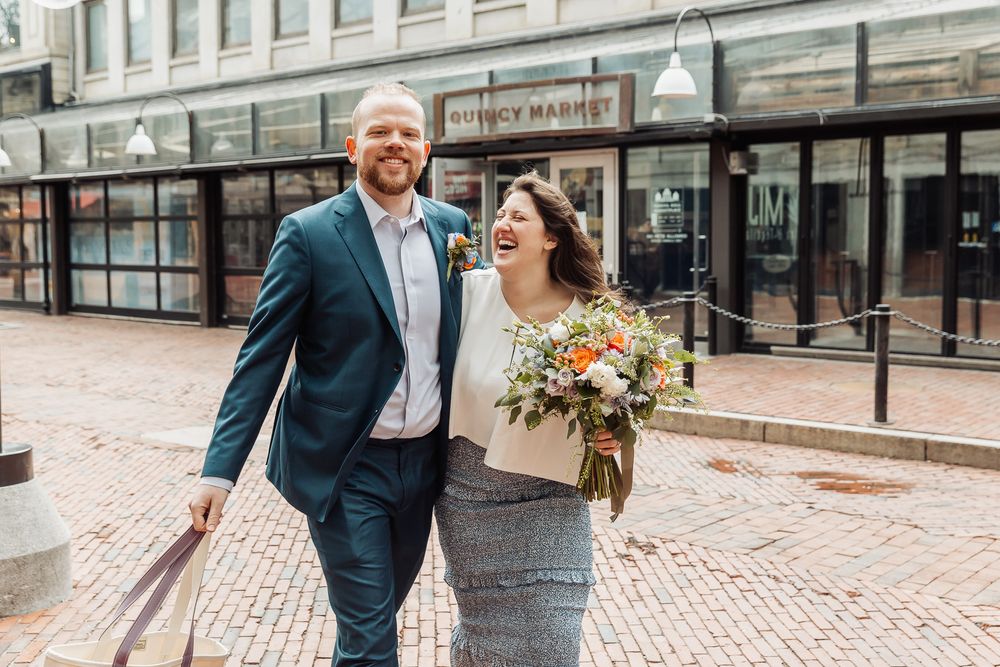 Happy couple laughing and walking together outside Quincy Market, bride holding colorful bouquet, groom in navy suit.