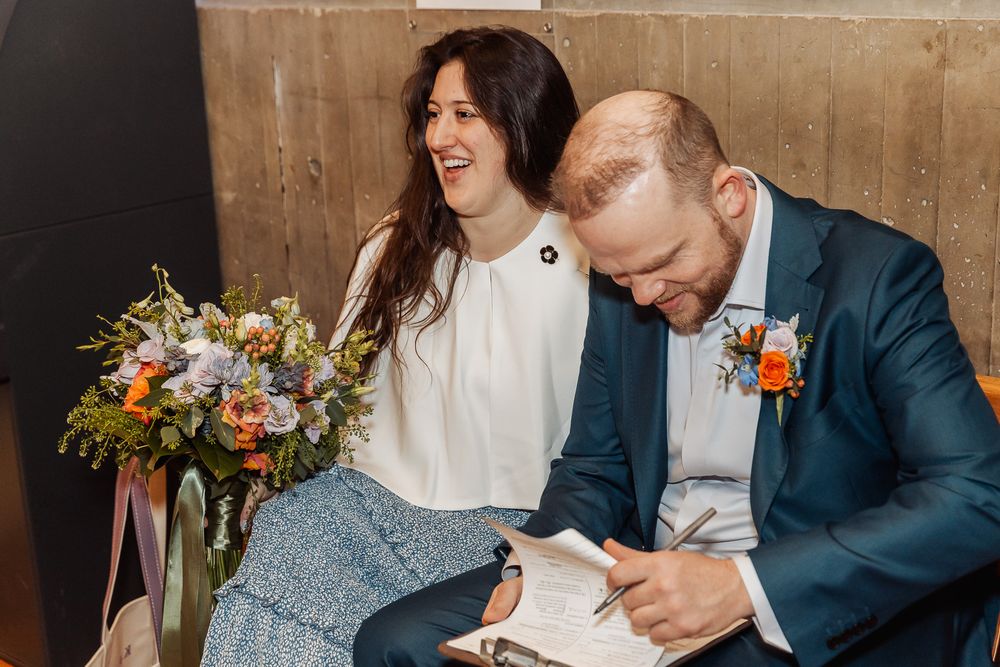 Bride laughing joyfully holding wildflower bouquet beside groom signing wedding register in navy suit with boutonniere.
