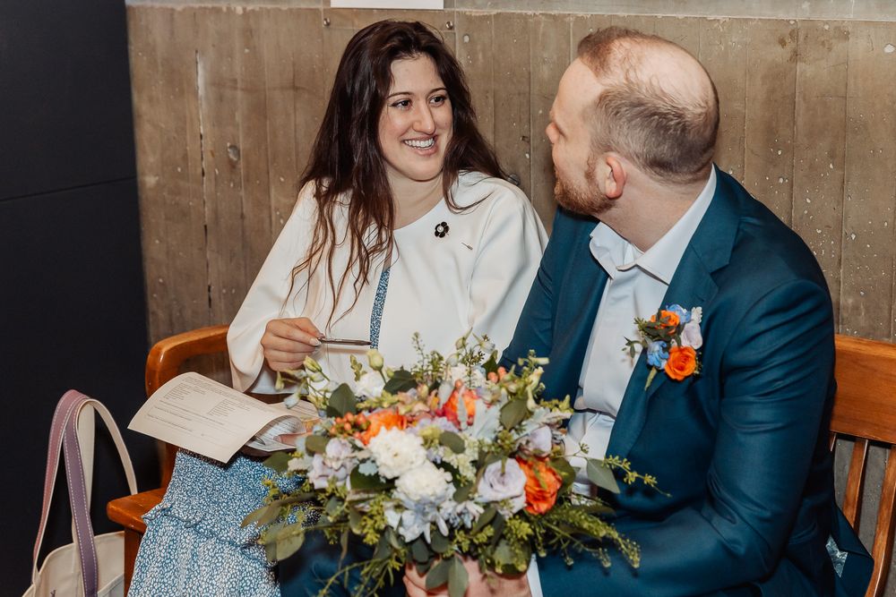 Smiling bride and groom sitting together indoors, bride holding colorful floral bouquet, groom in navy suit with boutonniere.