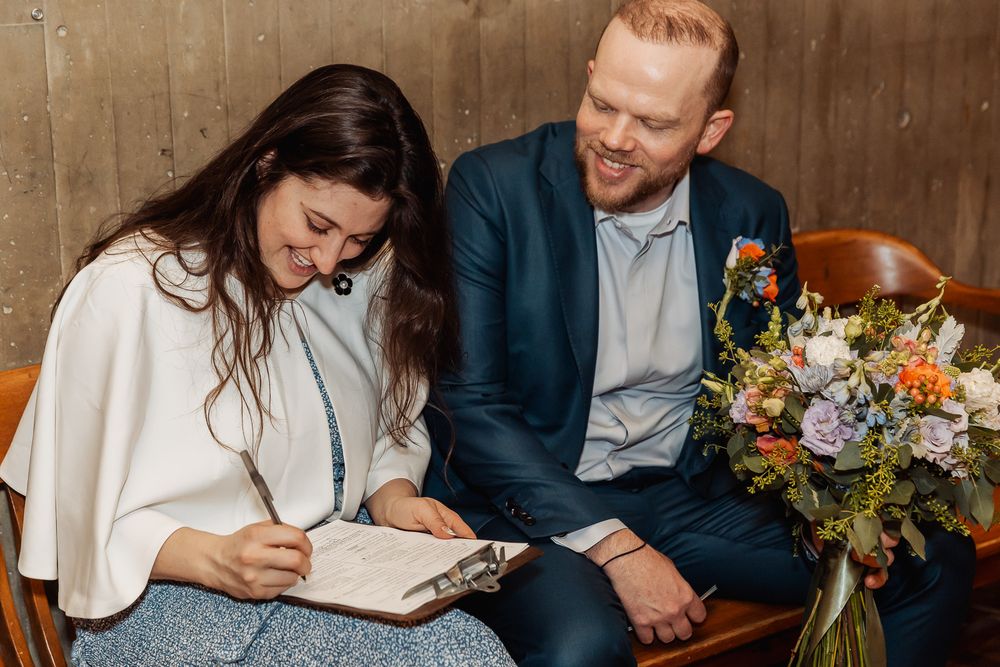 Smiling couple signing wedding documents, groom in navy suit holding colorful bridal bouquet indoors.