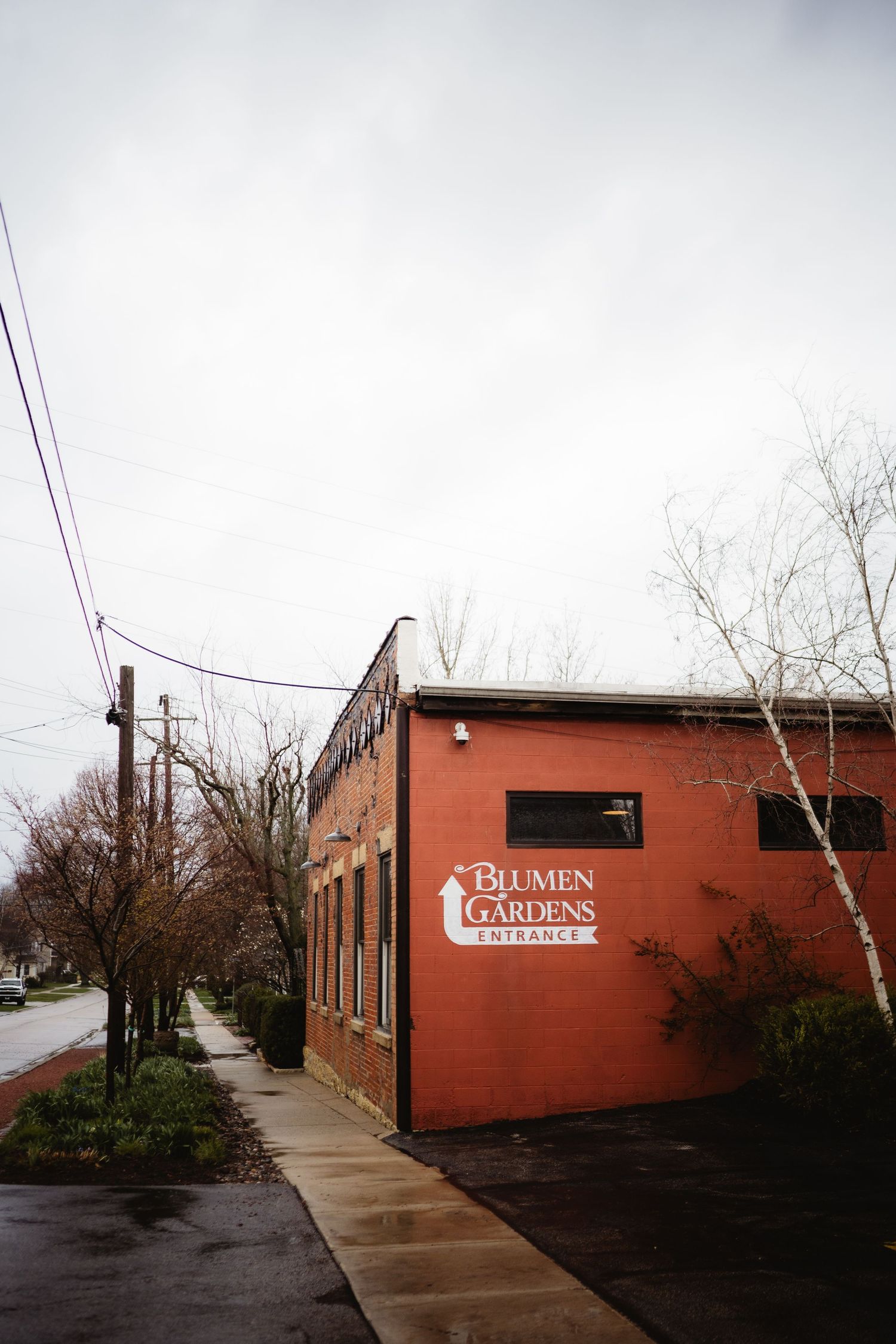 Red brick Blumen Gardens entrance building on a rainy day with bare trees and empty sidewalk.
