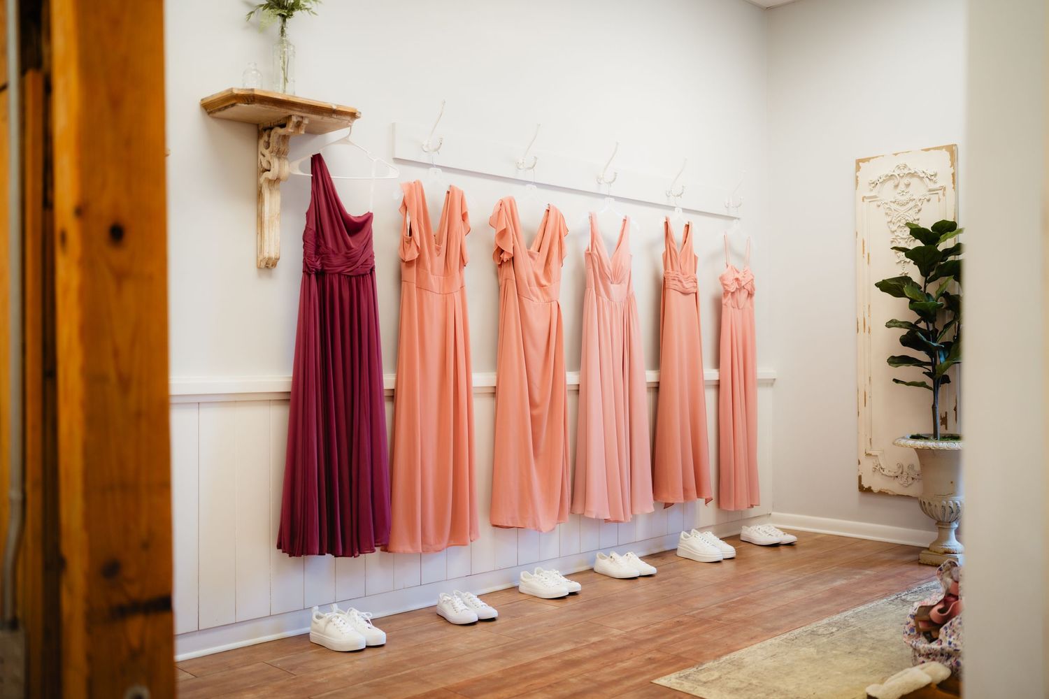Bridesmaid dresses in shades of burgundy and blush pink hanging on a rack with white heels below.