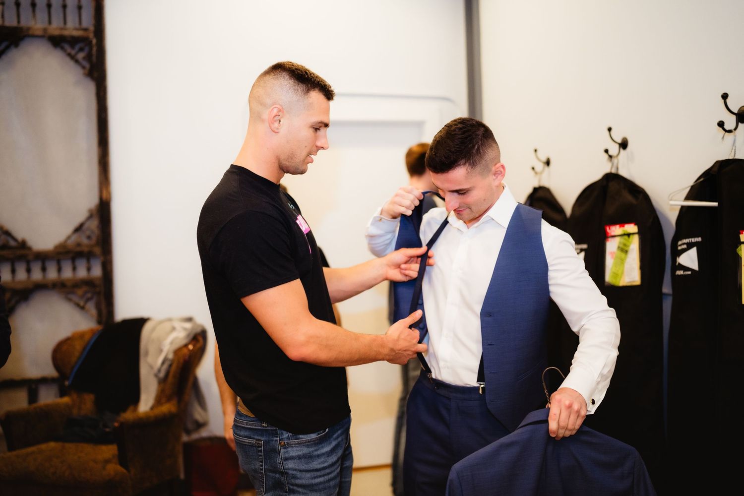 A tailor helps a groom adjust his blue vest and white dress shirt in a wedding preparation room.