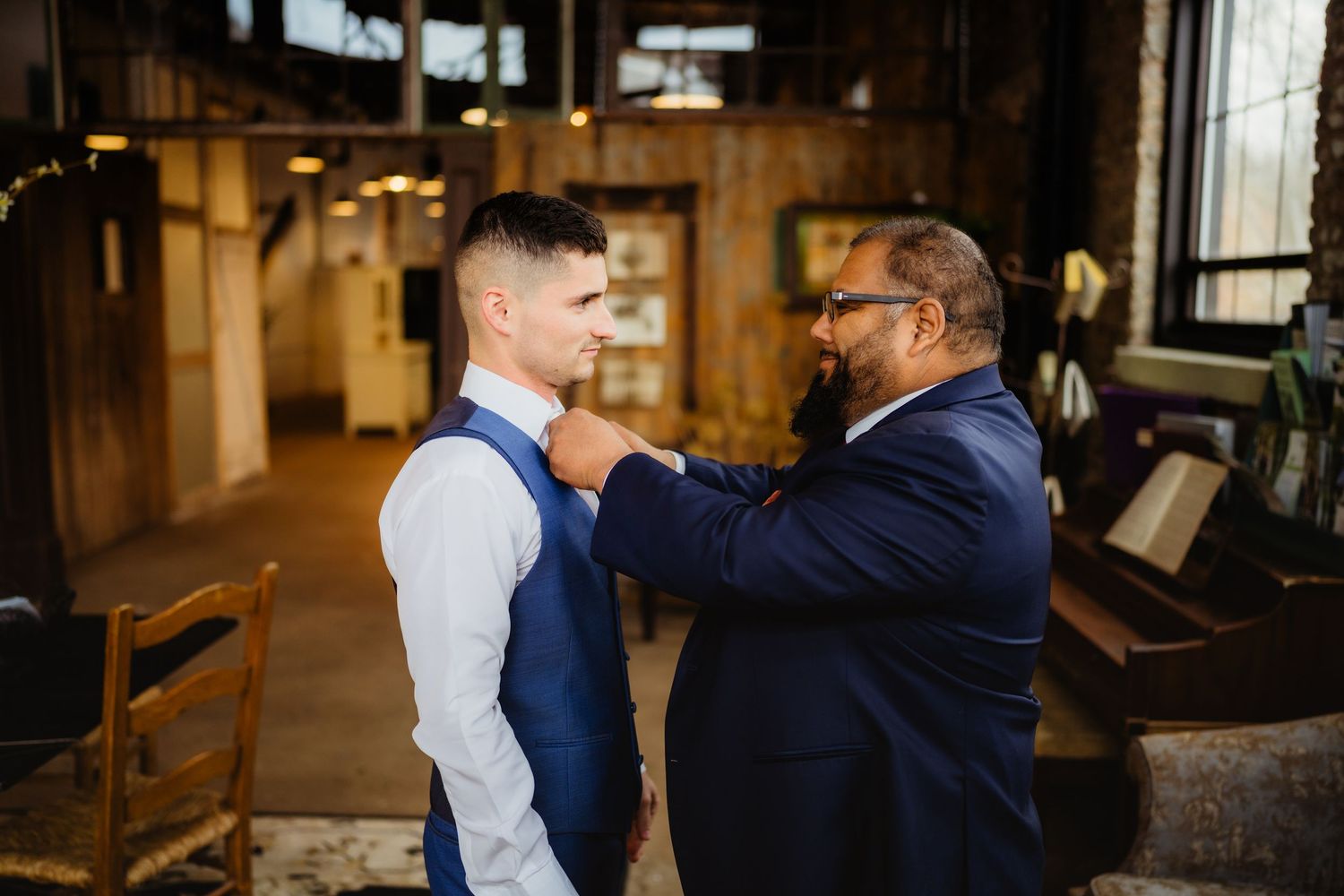 Man in navy suit adjusting boutonniere on groom in blue vest inside rustic wedding venue.