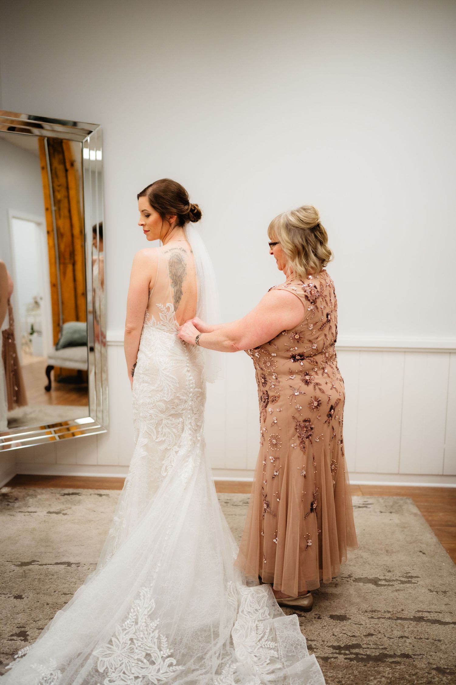 A bride in a lace mermaid gown gets her dress fastened by a woman in a rose gold beaded dress before her wedding.