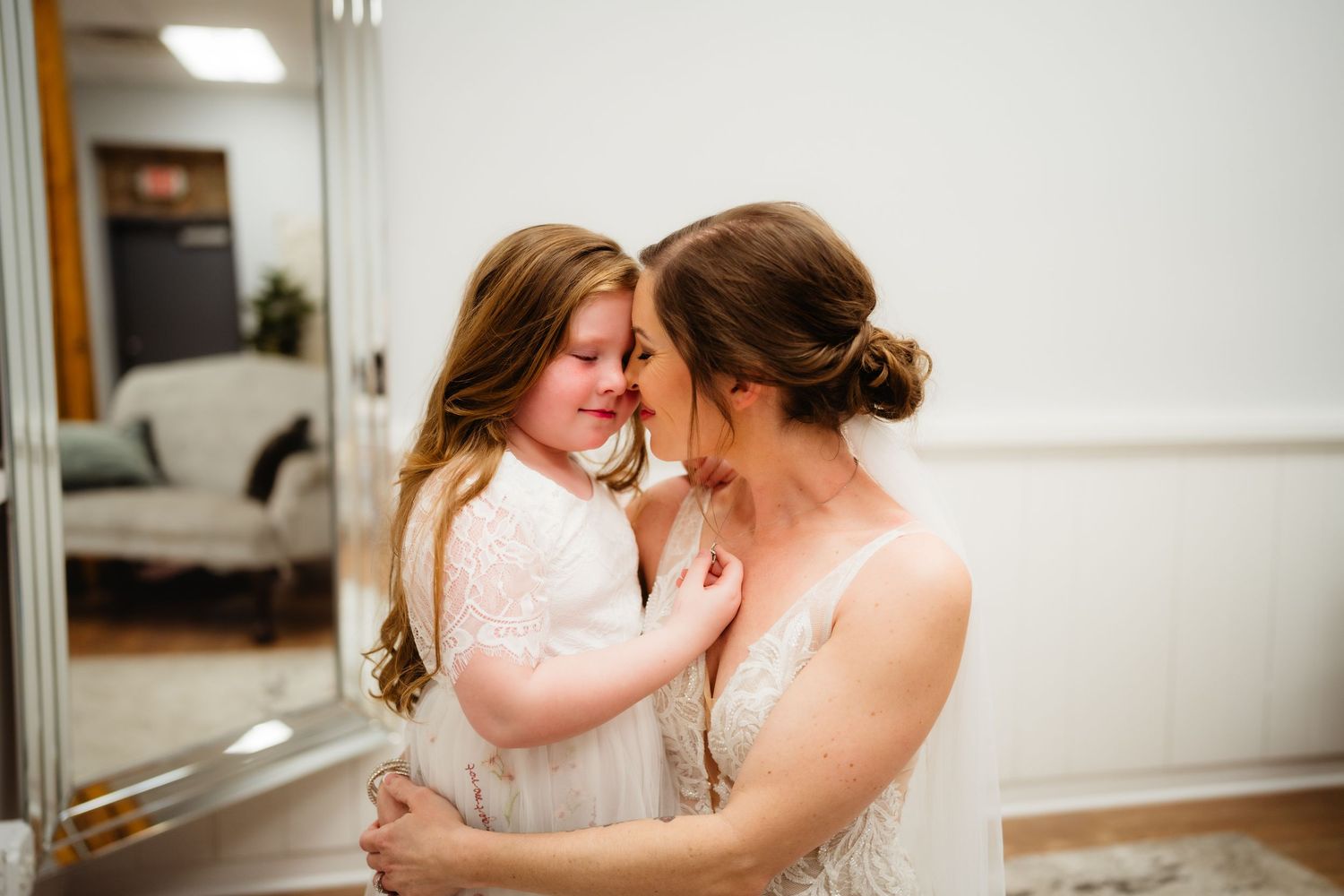 A bride in a white wedding gown shares a tender moment with a young flower girl in a white dress.