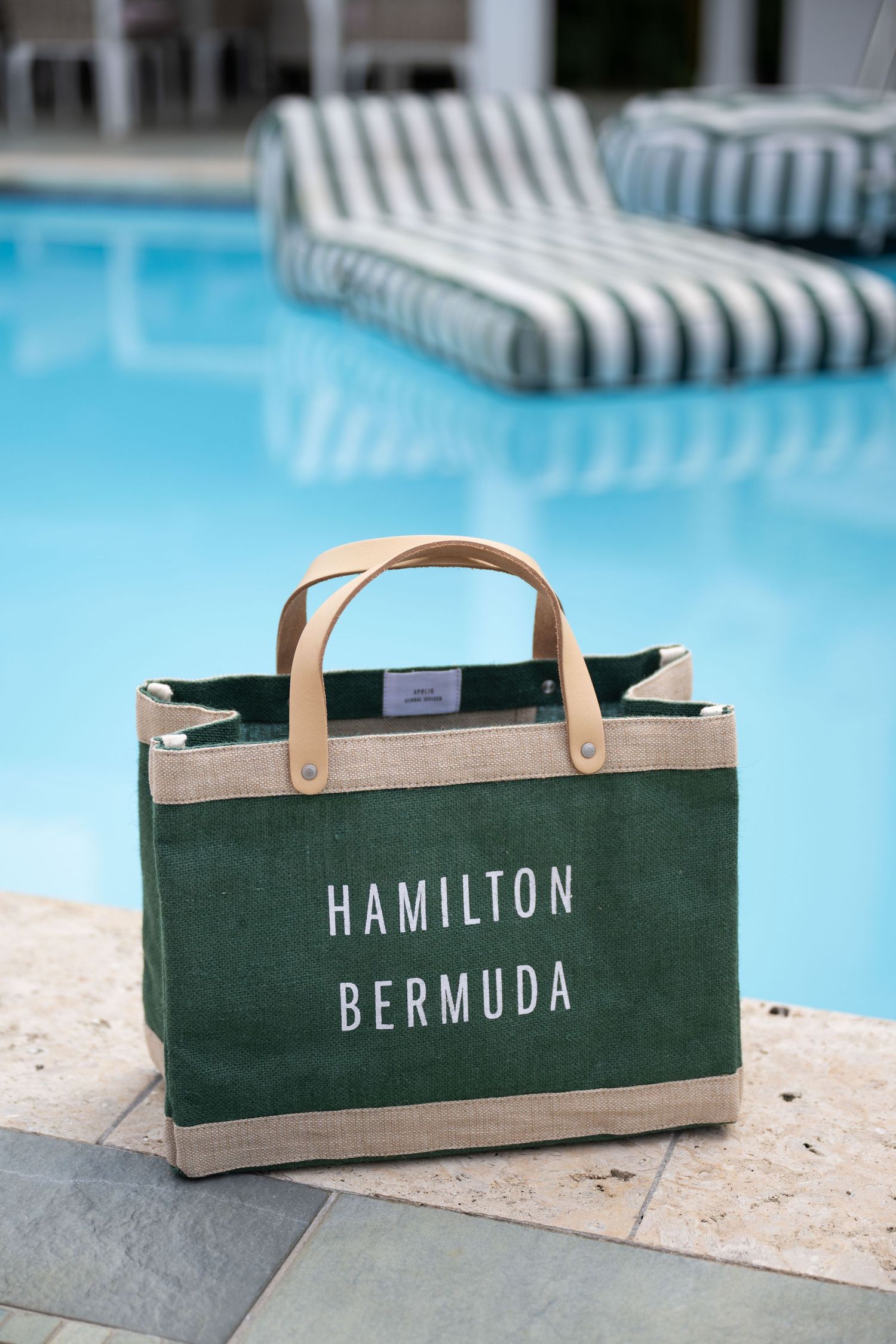 Green canvas tote bag with leather handles reading Hamilton Bermuda, placed poolside with striped lounge chairs.