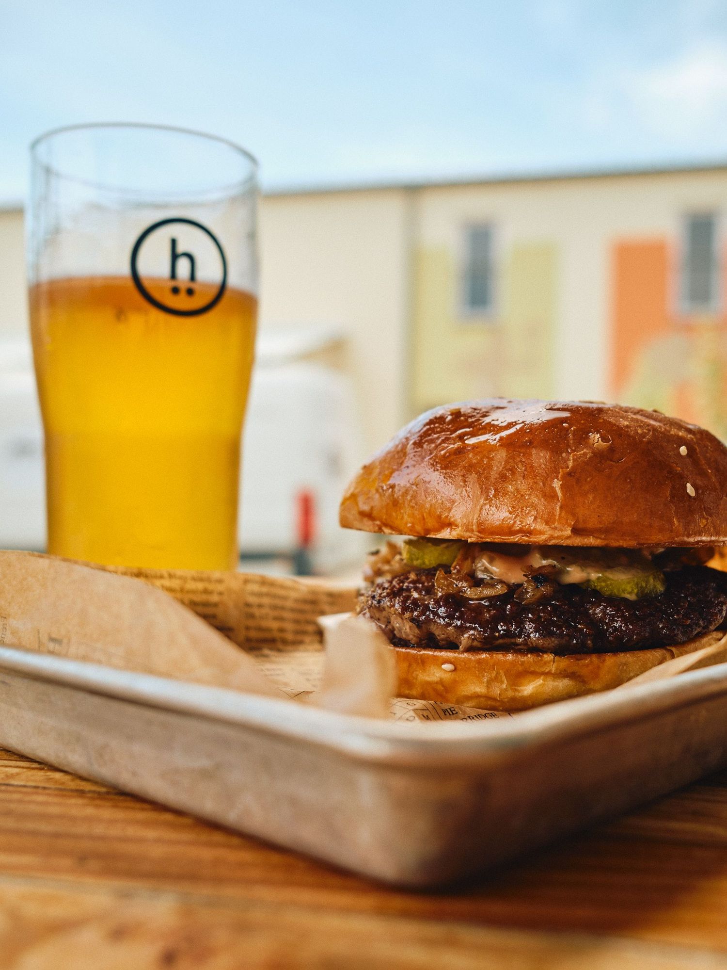 A juicy burger on a metal tray beside a cold pint of craft beer on a wooden table outdoors.