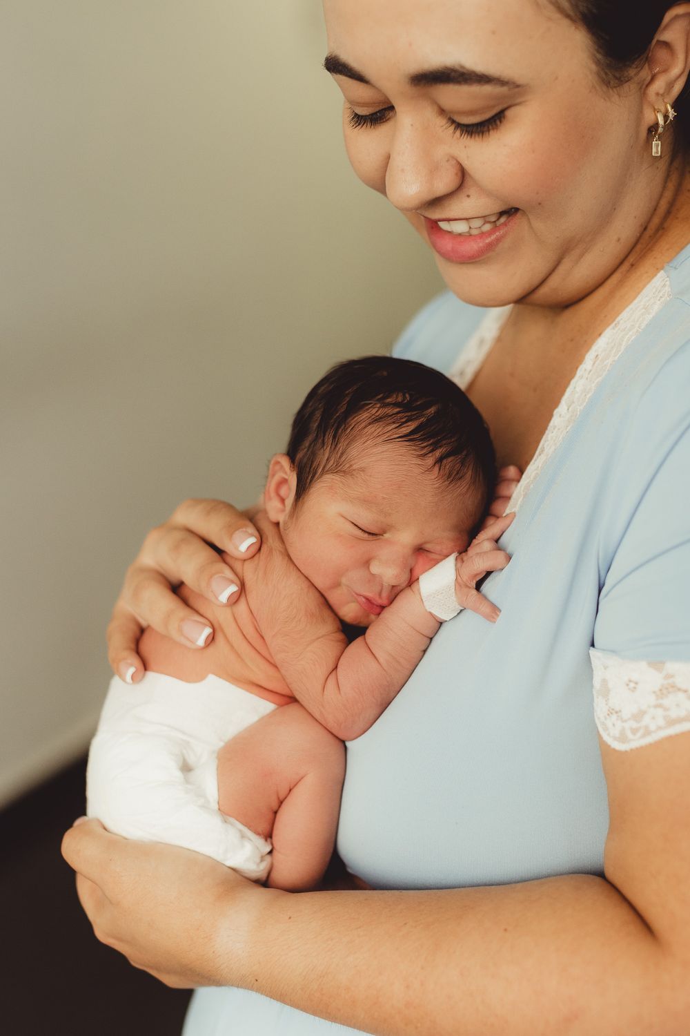 Smiling mother in blue dress gently holding her sleeping newborn baby wrapped in white cloth.