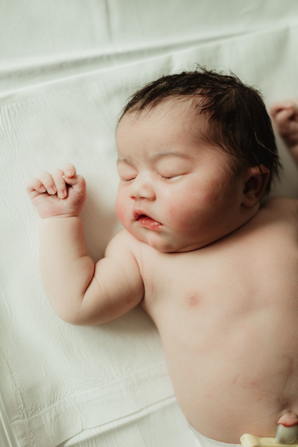 Peaceful newborn baby sleeping on white blanket with dark hair and tiny fist raised.