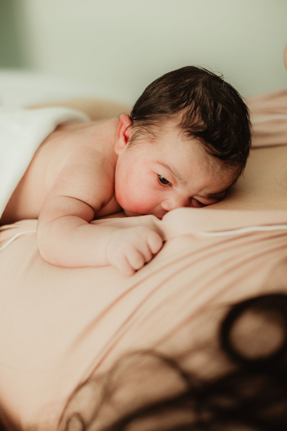 Newborn baby with dark hair lying on a peach blanket, eyes open, in a peaceful and tender moment.