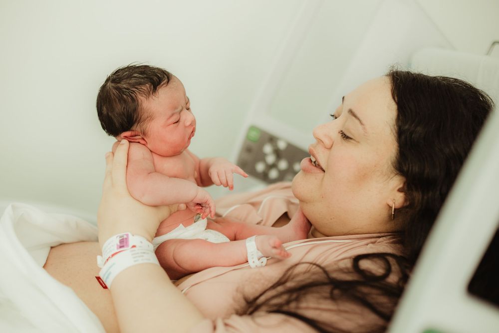 A new mother lying in a hospital bed smiles lovingly at her newborn baby during skin-to-skin contact after birth.