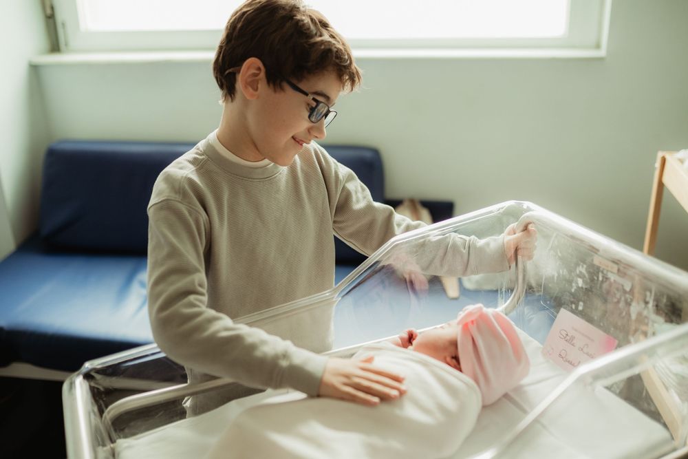 Young boy with glasses gently touching a newborn baby in a hospital bassinet.