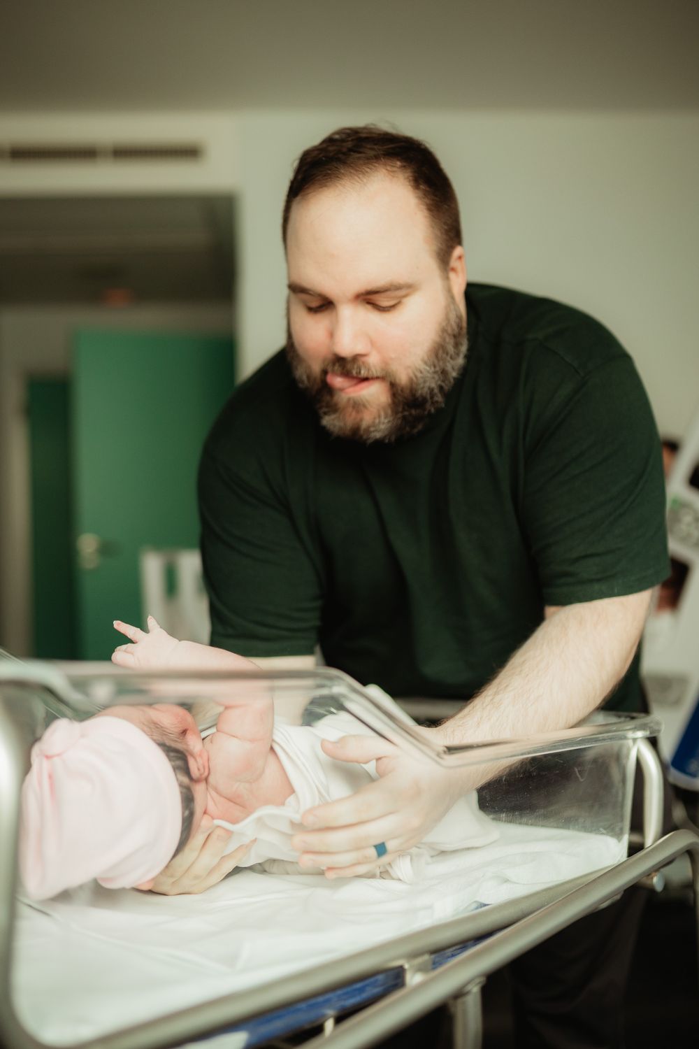 Bearded father in green shirt tenderly looks at newborn baby in hospital bassinet.