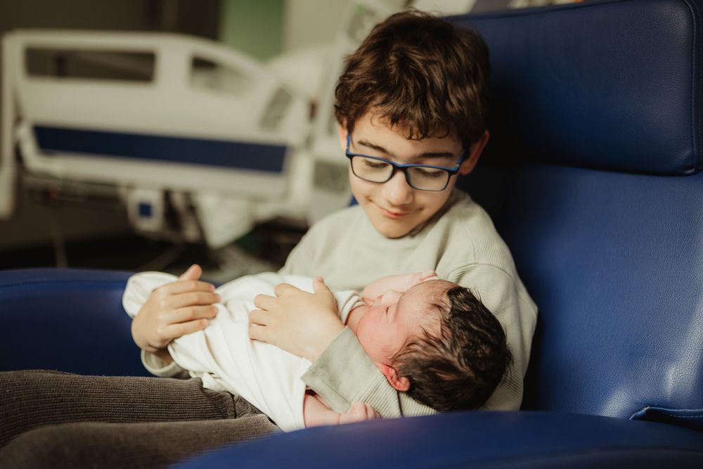 Young boy with glasses holding a newborn baby sibling in a hospital room chair.