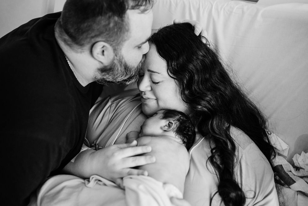 Black and white photo of parents lovingly kissing their newborn baby together.