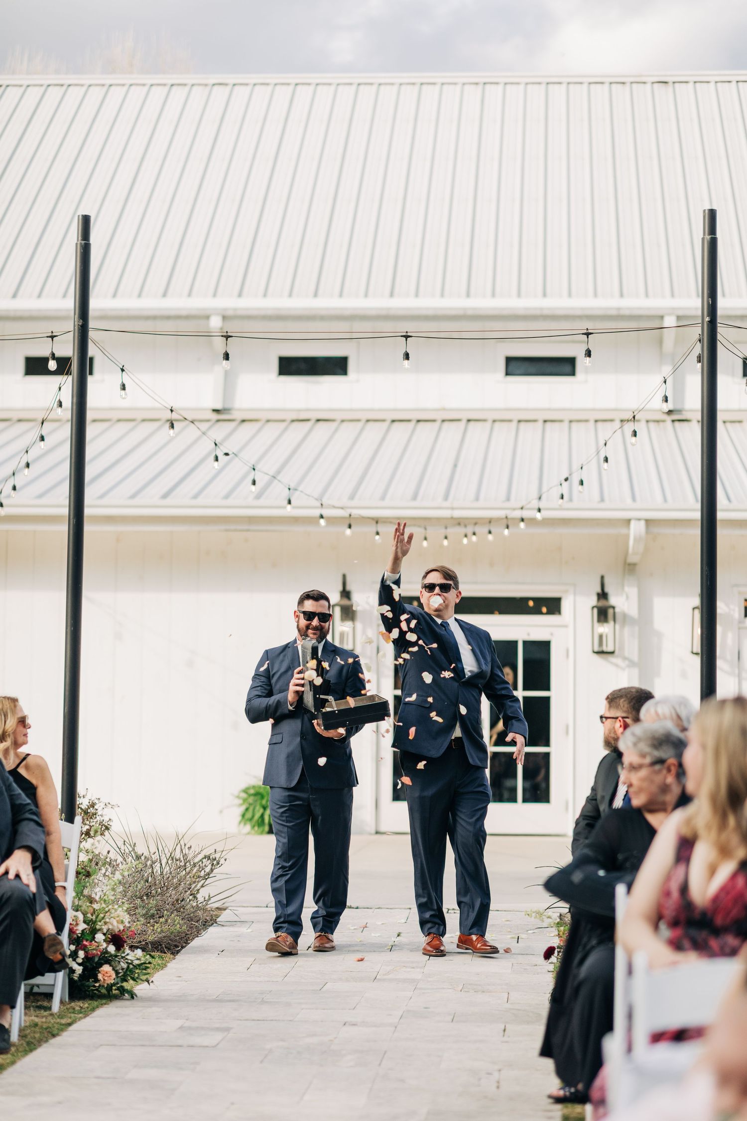 Wedding party members in dark suits playfully posing with toy guns under a white covered patio.