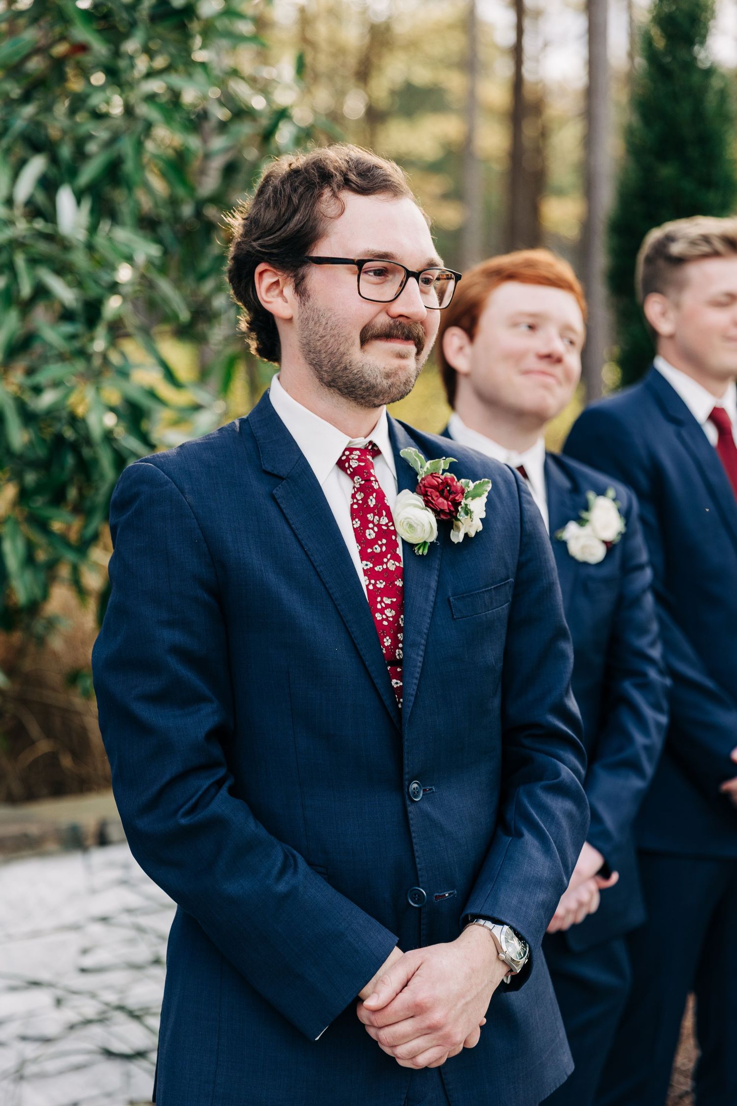 Groomsmen in navy suits and red ties standing together at an outdoor wedding ceremony.