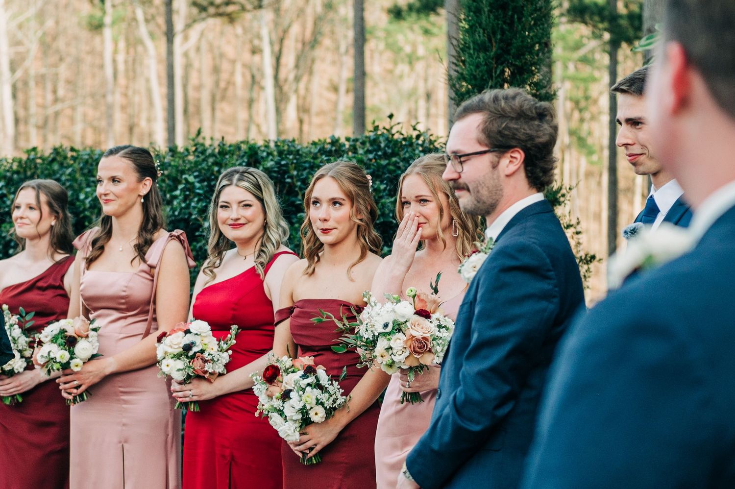 Wedding party members in burgundy and pink dresses pose with white floral bouquets in an outdoor setting.