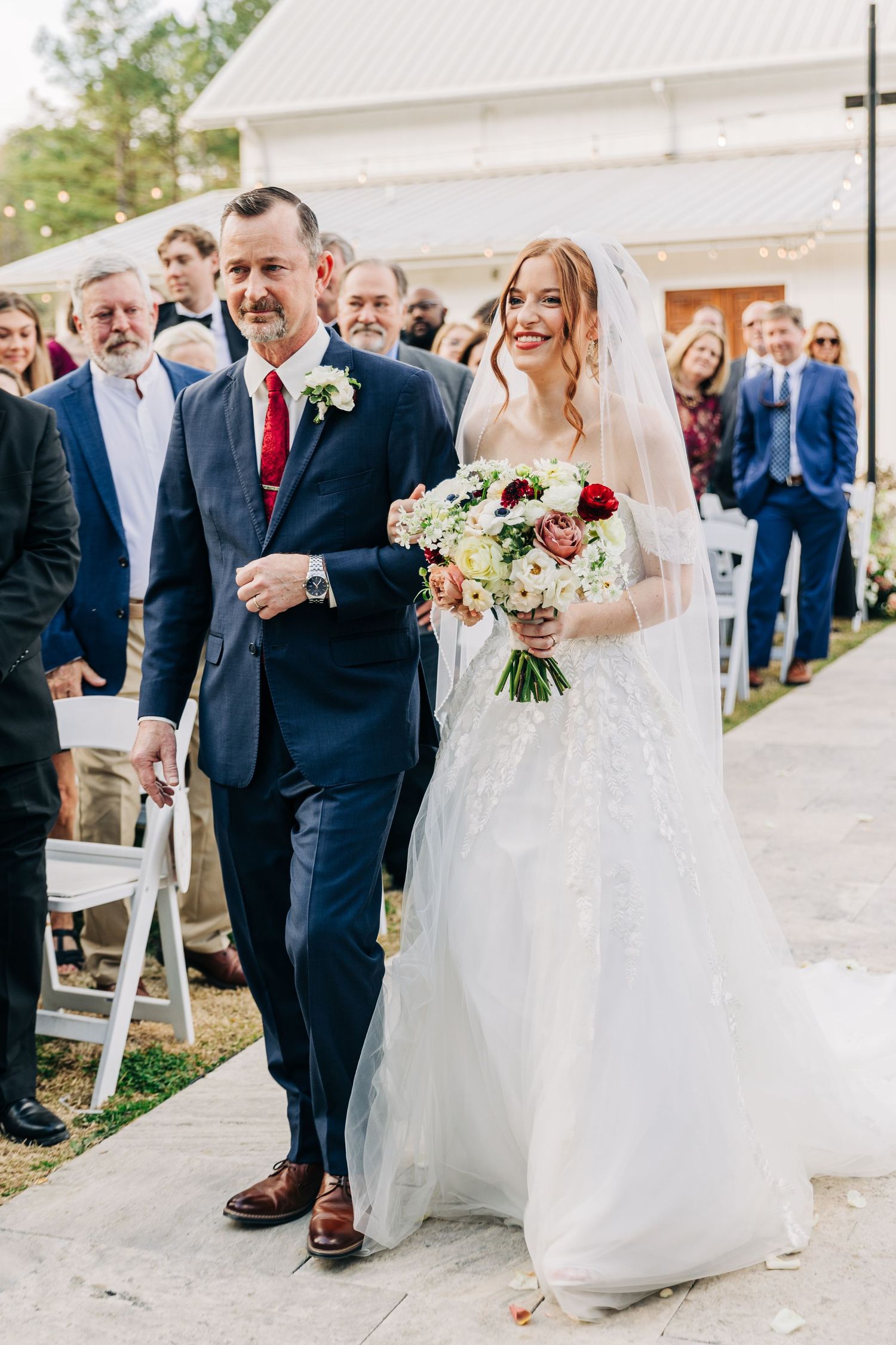 A wedding ceremony processional showing a white dress and navy suits against an outdoor backdrop with guests watching.