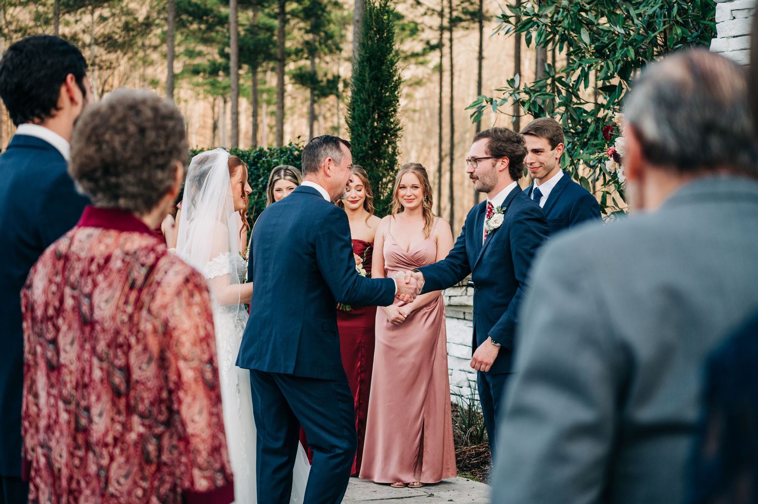 Wedding guests and bridal party gathered outdoors with greenery and trees in the background.