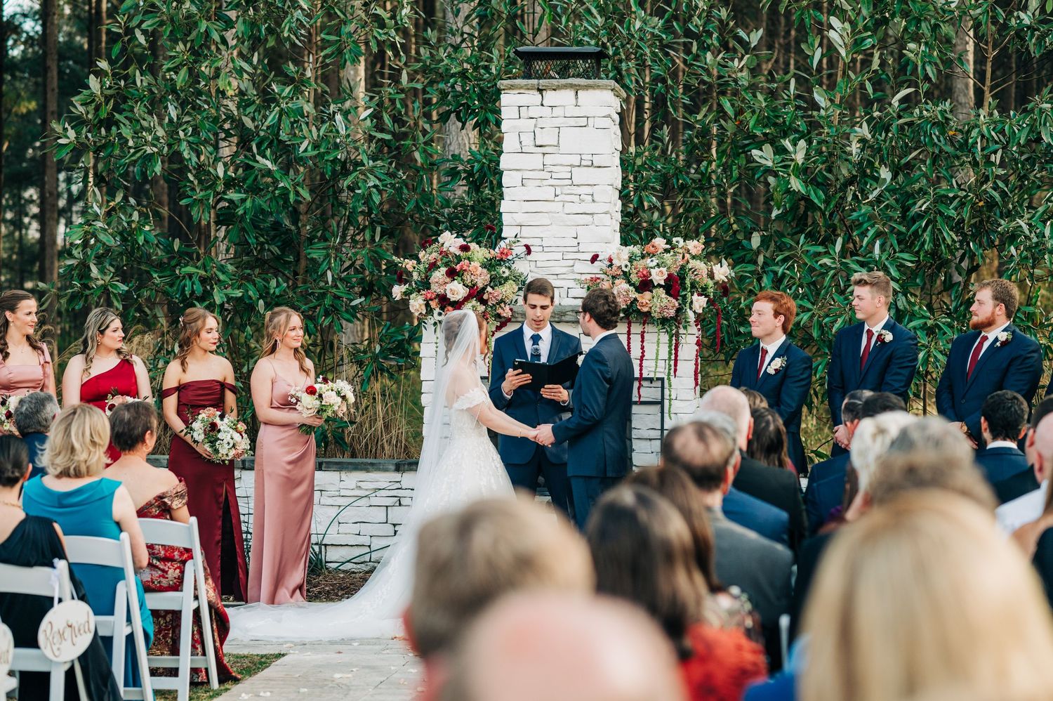 Outdoor wedding ceremony in a lush garden setting with guests seated in white chairs facing a white brick wall.