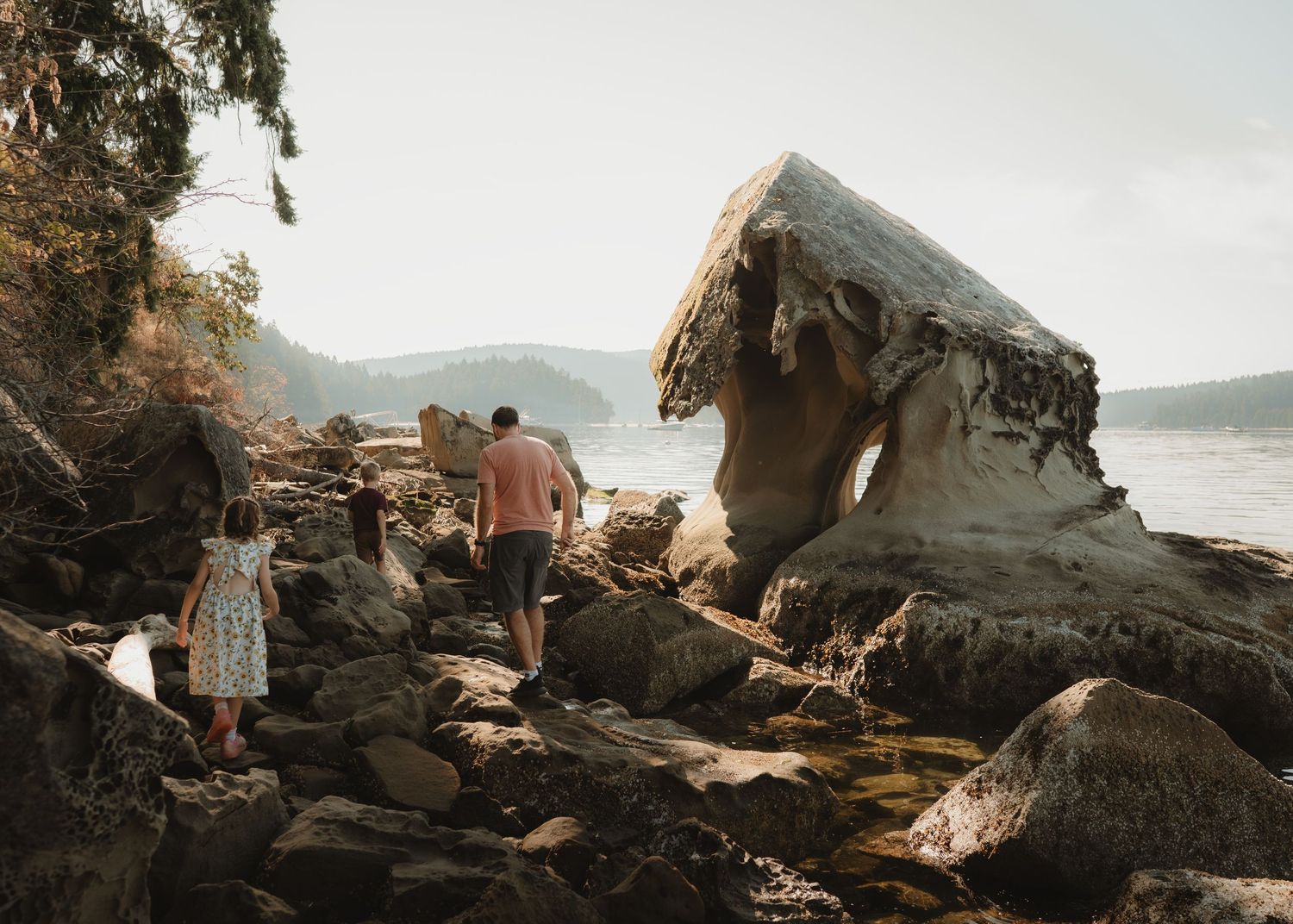 Two people explore a rocky shoreline beside a uniquely shaped sandstone rock formation with calm water and hills in the background.