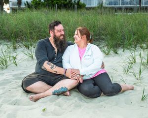Couple sitting together on sandy beach, holding hands and smiling, with dune grass in background.