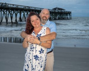 Couple embracing and smiling on a sandy beach with a pier in the background at dusk.