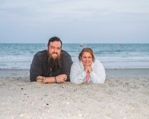 Couple lying on sandy beach smiling, man with beard and tattoos, woman in white jacket, ocean in background.