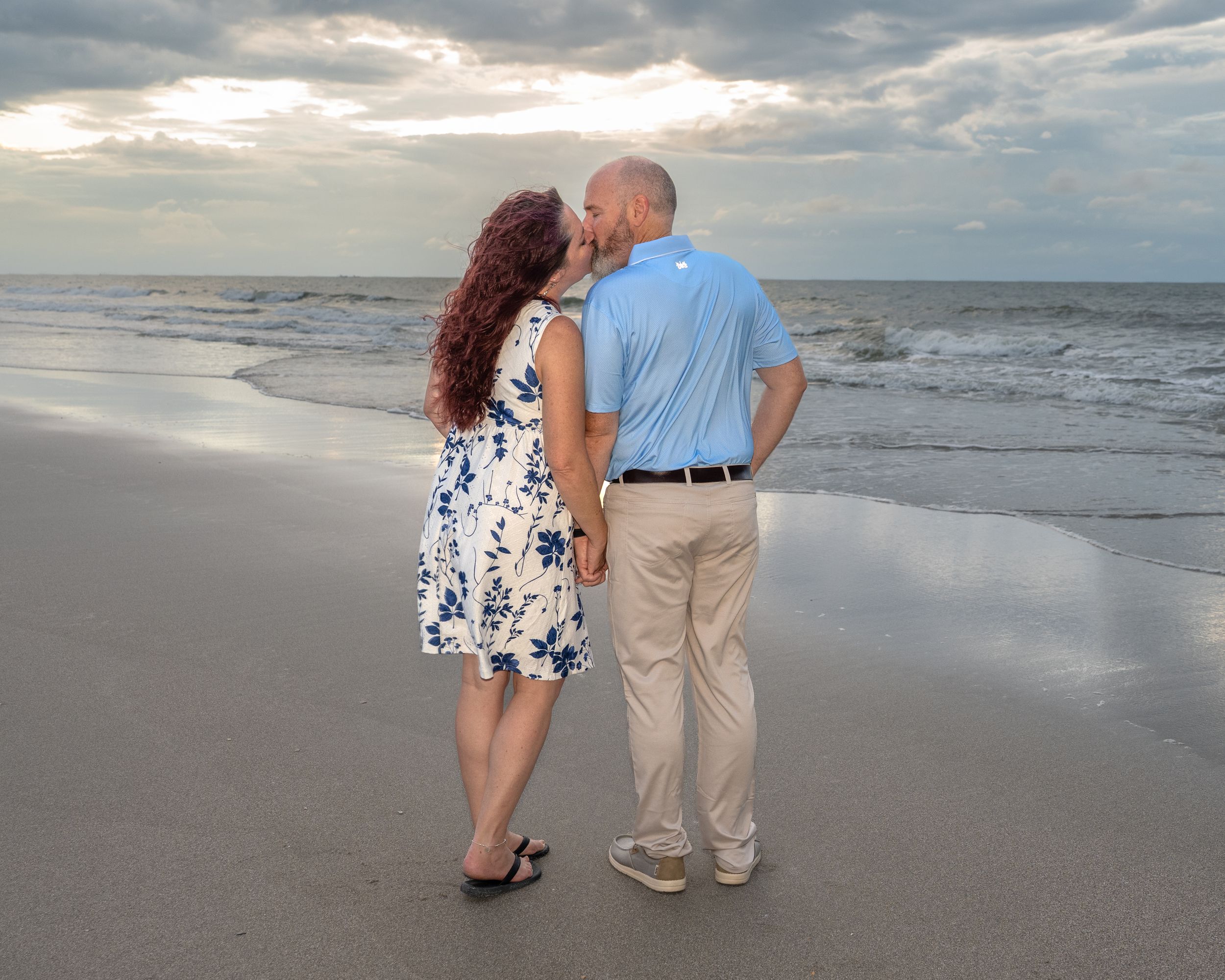 Couple sharing a romantic kiss on a sandy beach at sunset with ocean waves in the background.
