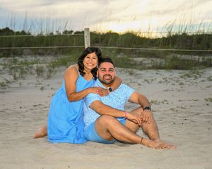 Couple in matching blue outfits sitting on sandy beach at sunset, smiling and embracing near dune fence.