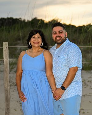 Couple in matching blue outfits holding hands on a sandy beach path at sunset with dunes in background.