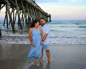 Couple holding hands smiling on beach at sunset near pier, woman in blue dress, man in floral shirt.