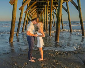Couple sharing a romantic kiss under a wooden beach pier at golden sunset with ocean waves in the background.