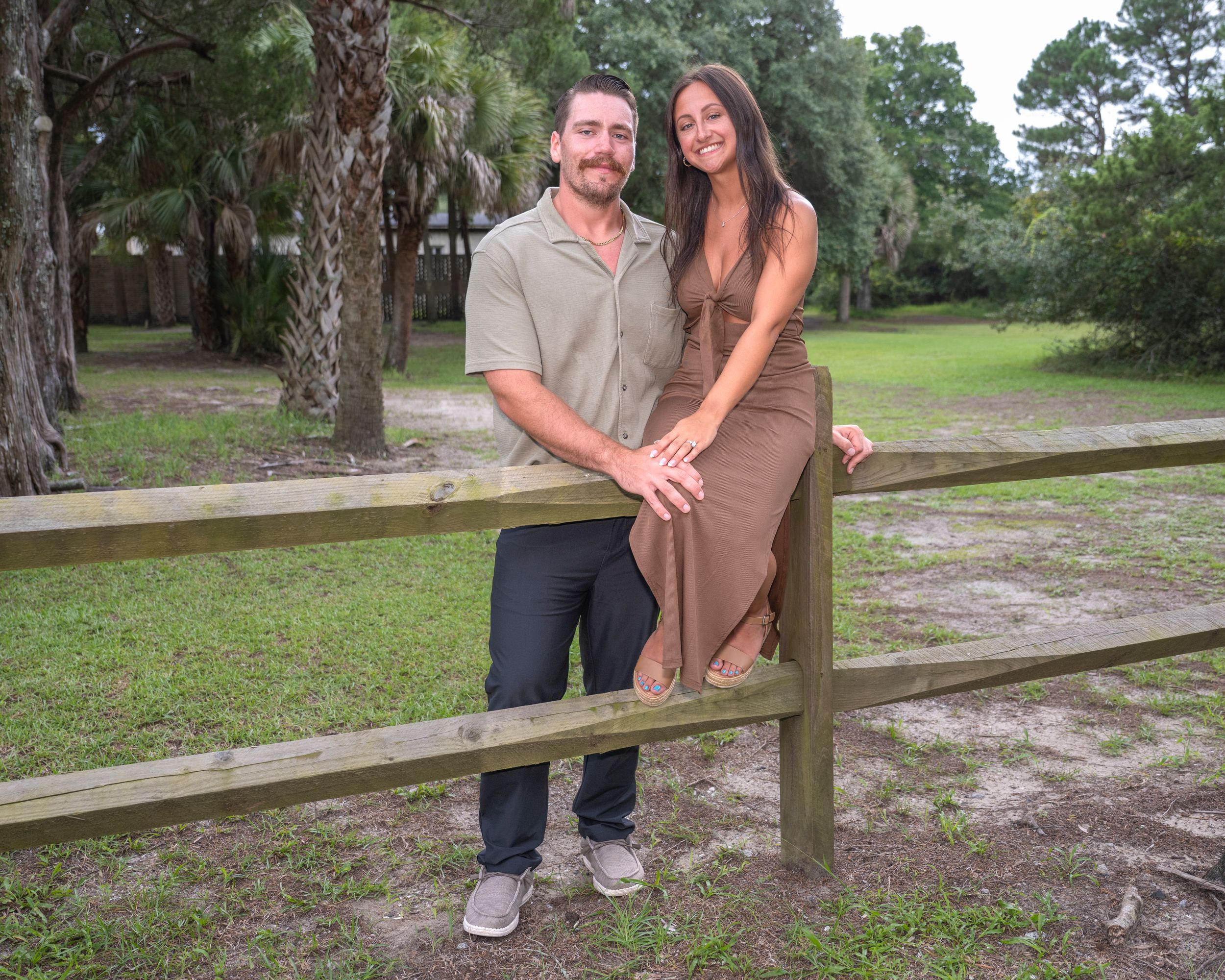 Smiling couple posing together against a wooden fence in a lush green park setting.