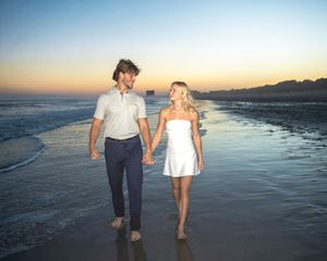 Couple holding hands walking on beach at sunset, man in polo and navy pants, woman in white dress.