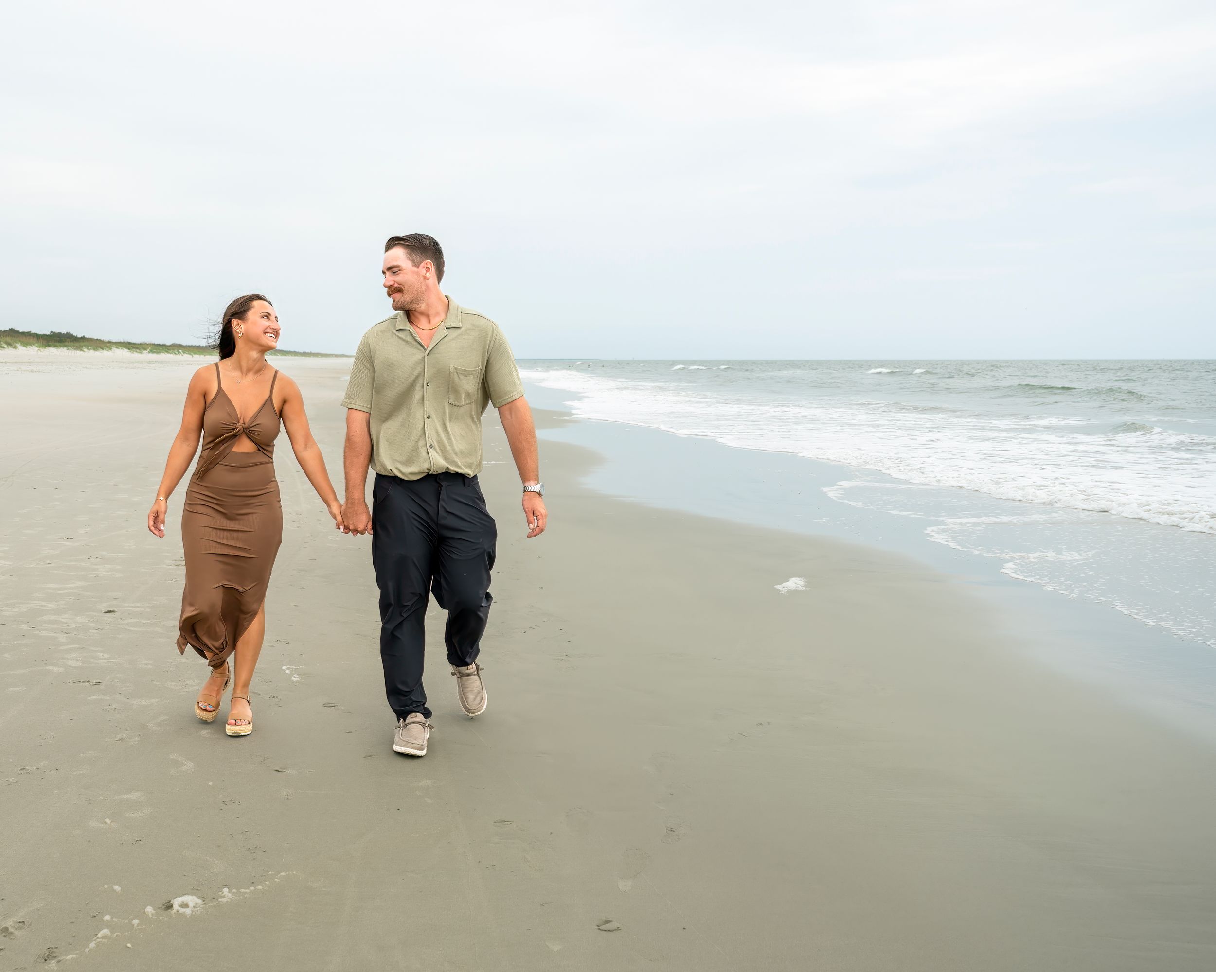 Couple holding hands and walking together on a sandy beach near ocean waves on a cloudy day.