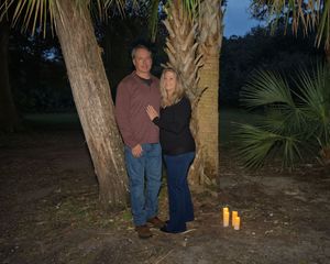 Couple standing together by palm trees at night with candles on the ground in an outdoor setting.