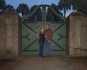 Couple standing together in front of large green wooden gates flanked by brick walls and palm trees at dusk.
