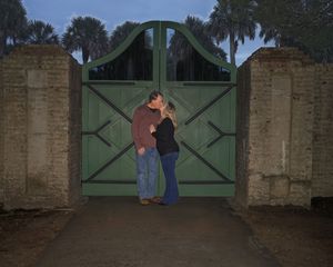 Couple kissing in front of a green wooden gate flanked by brick pillars and palm trees at dusk.