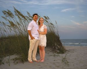 Romantic couple in white clothing standing on coastal sand dunes with sea grasses and ocean waves in background.