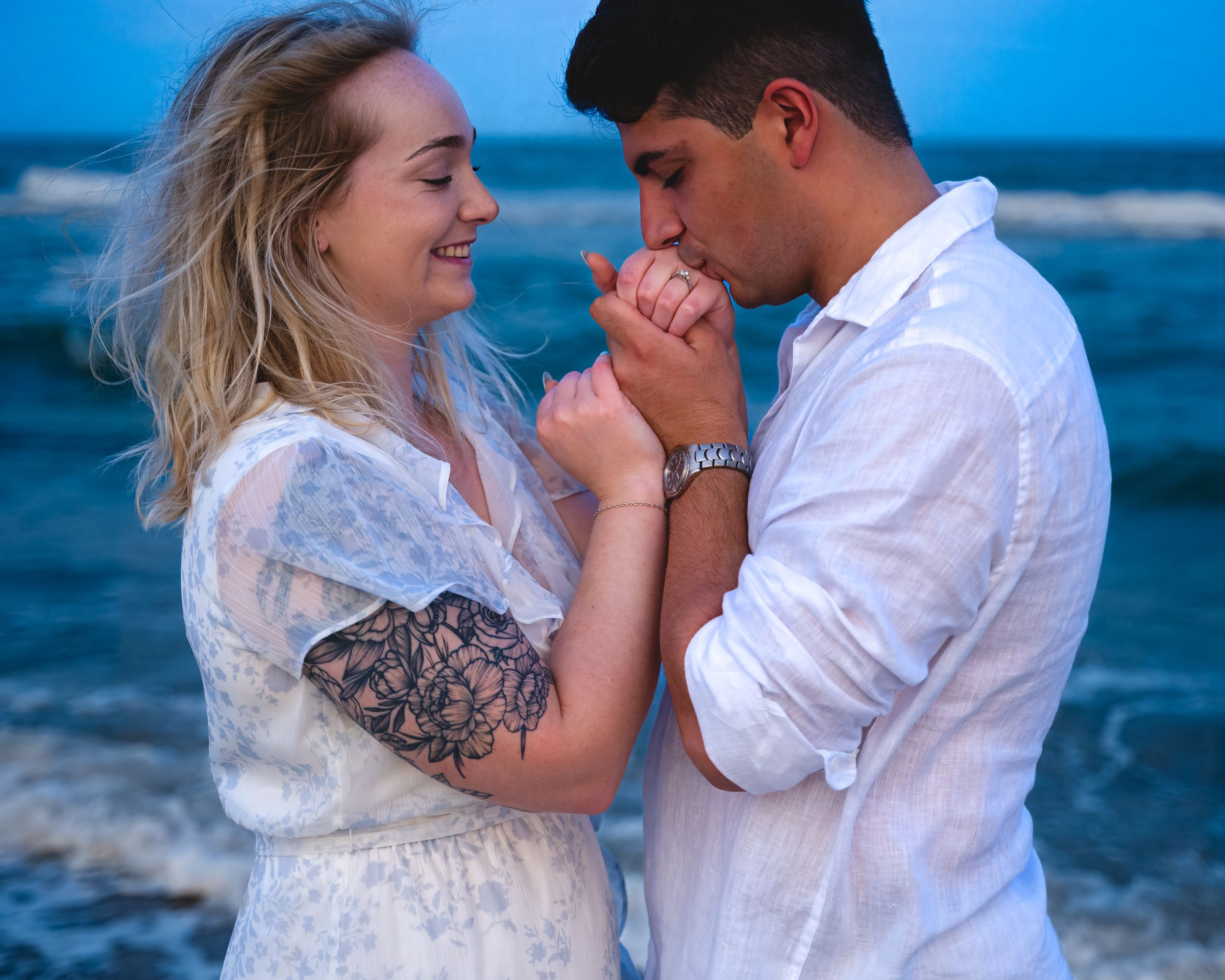 A couple shares romantic moments in white outfits while standing along a scenic ocean shoreline at sunset.