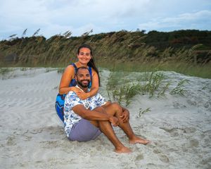 Two people enjoying a relaxing moment together on a sandy beach with sea oats and dunes in the background.