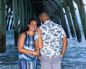 A couple in blue and floral patterns stand underneath a wooden pier near ocean waves on a bright day.