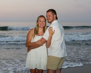 A couple shares a romantic moment on a beach at sunset while waves crash behind them in the background.