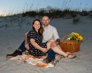 A couple enjoys a romantic beach picnic at dusk with a wicker basket and sunflowers on sandy shores.