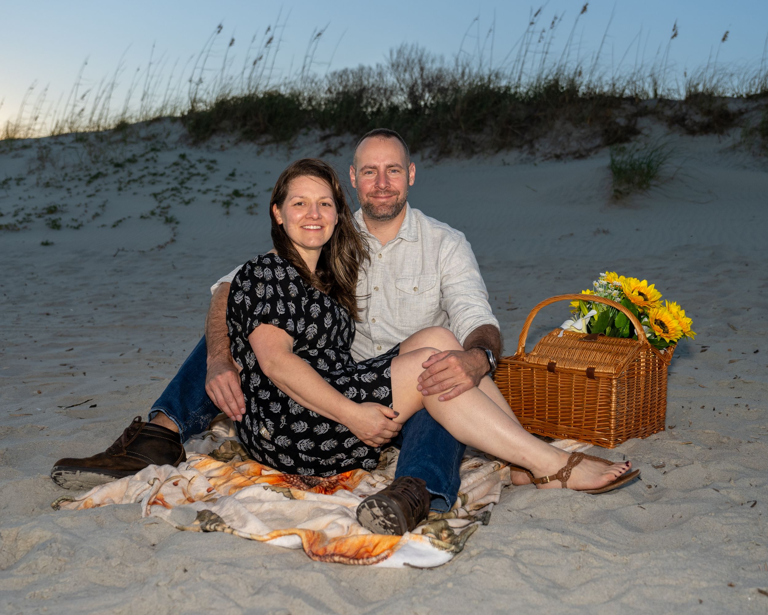 A couple enjoys a romantic beach picnic at dusk with a wicker basket and sunflowers on sandy shores.