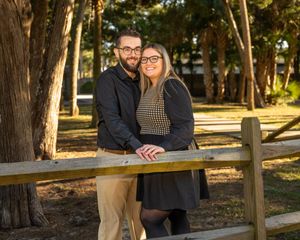 A romantic couple poses together outdoors along a wooden fence in a sunlit forest setting.